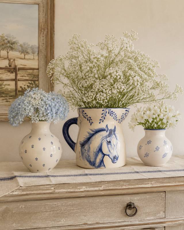 Jarra de cerâmica com cavalo azul pintado à mão, com flores brancas, sobre uma cômoda