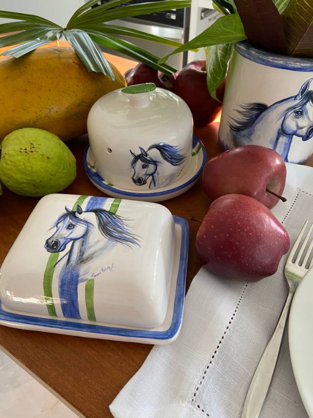 Manteigueira de cerâmica com cavalo azul e listras verdes e azuis, ao lado de frutas