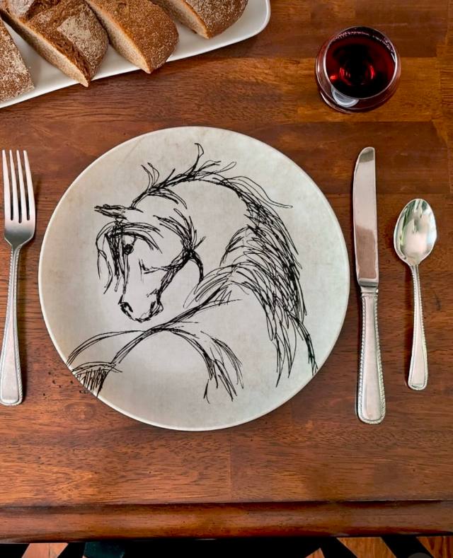 Prato de cerâmica com cavalo em traços pretos, em mesa com pães e vinho