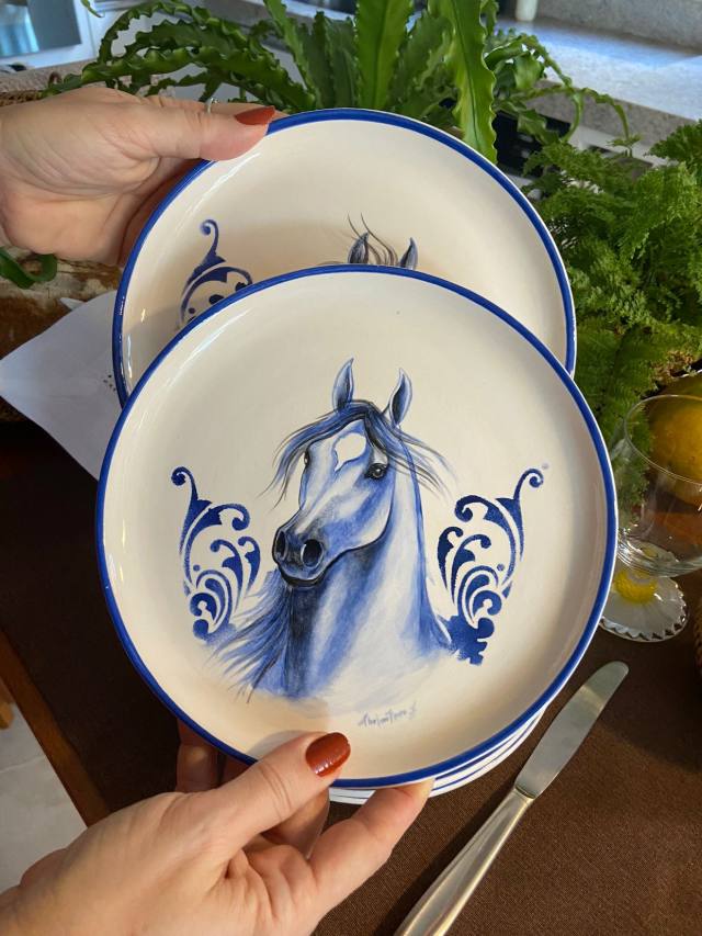 Mãos segurando pratos de cerâmica com cavalo azul e arabescos pintados à mão