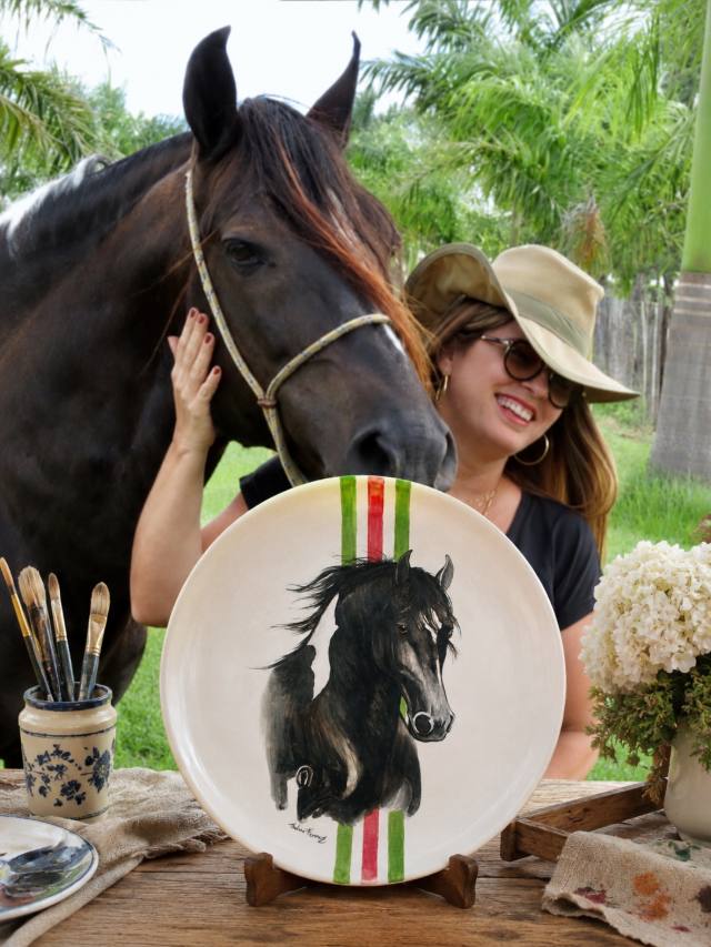Mulher abraçada a um cavalo preto, com o retrato do animal pintado em um prato de cerâmica em primeiro plano