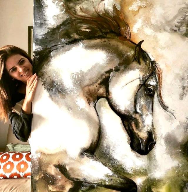 Mulher sorrindo ao lado de uma tela grande com cavalo tordilho em tons de ocre