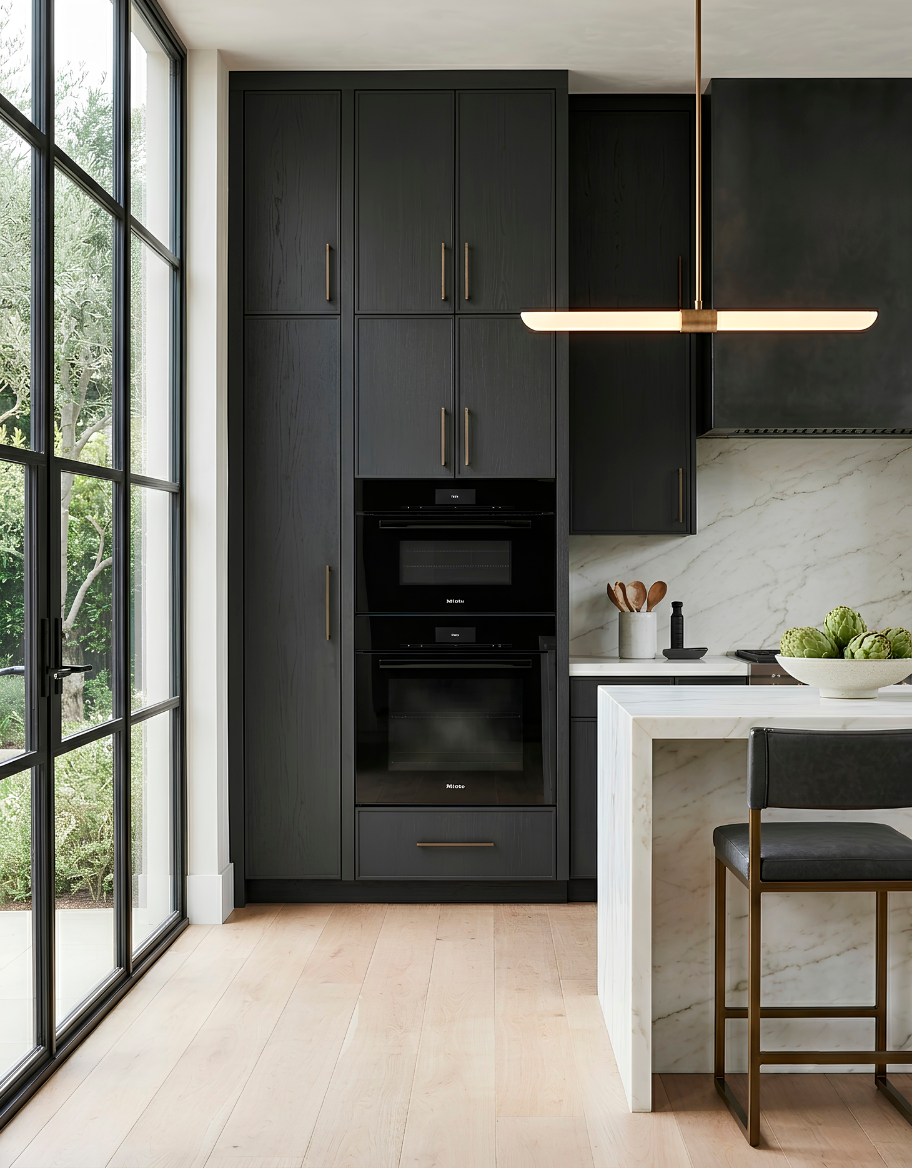 Dise&ntilde;o de cocina moderna de lujo con mueble torre de hornos en carpinter&iacute;a negra, isla central de m&aacute;rmol blanco Calacatta y ventanal de hierro negro.
