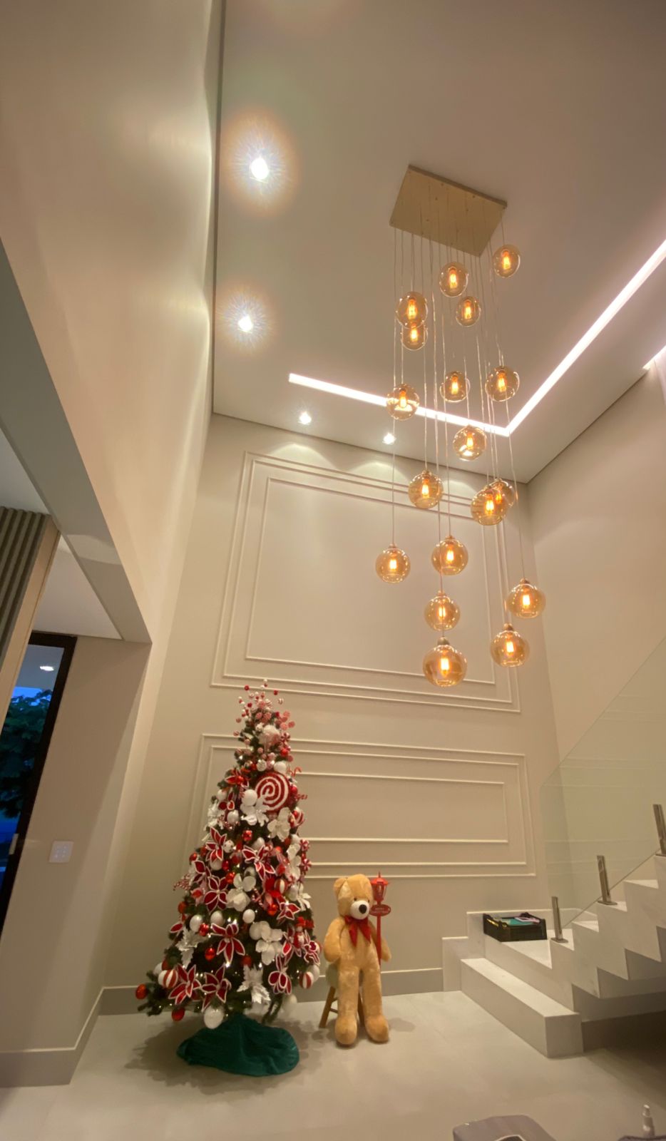 Sala com lustre pendente de esferas âmbar, árvore de Natal decorada e urso de pelúcia ao lado da escada.