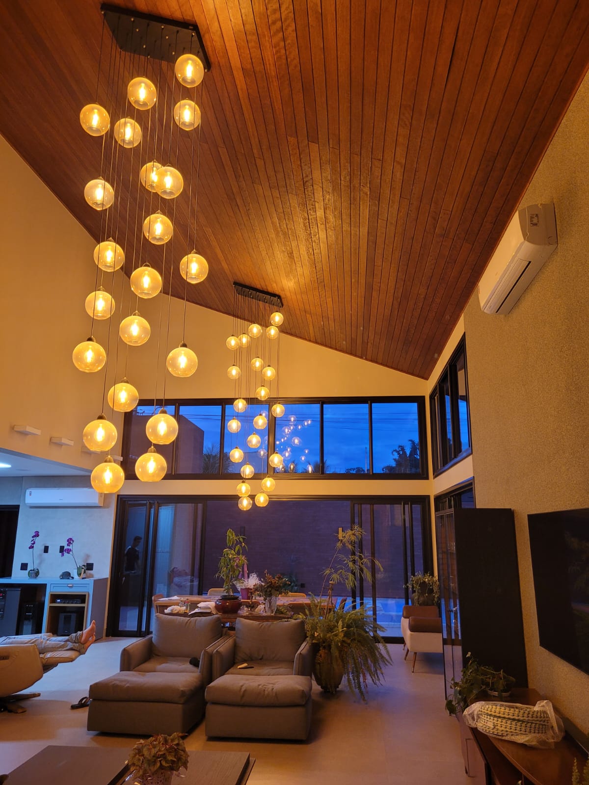 Sala de estar ampla com teto de madeira inclinado, iluminada por lustres pendentes com esferas de vidro âmbar e janelas de vidro destacando a paisagem externa.