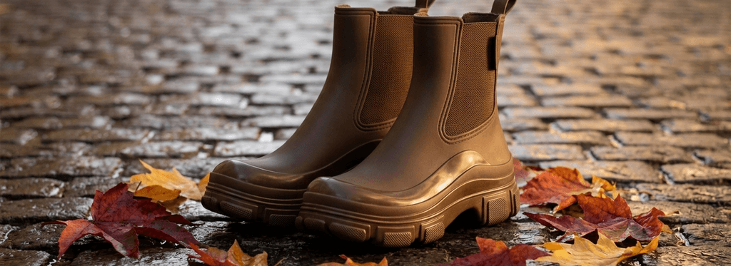 Par de botas Rain Valky chocolate sobre adoquines mojados con hojas de otoño, luz cálida al ocaso