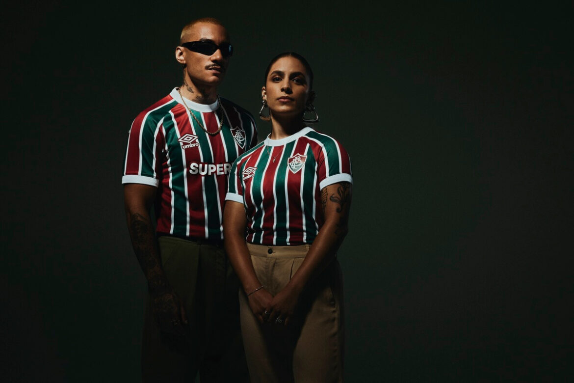 "Dois torcedores vestindo a nova camisa tricolor do Fluminense 2025/26 posam lado a lado em frente a um fundo escuro. As camisas apresentam listras verticais nas cores verde, grená e branco, com o escudo do Fluminense à direita e o logo da Umbro à esquerda, acompanhado da palavra 'SUPER' em branco logo abaixo. Os detalhes contrastantes da camisa se destacam na imagem, transmitindo identidade e paixão pelo clube."