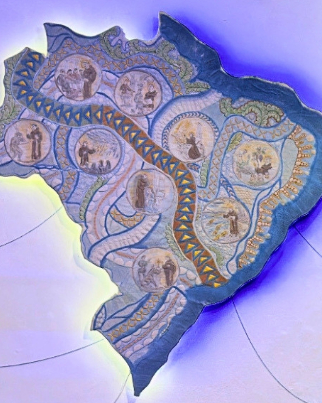 Fotografia em plano detalhe da obra de arte têxtil "Caminhos de Fé", de Hélen Jordana, destacando as pinturas em aquarela e os relevos do quilting. A peça em formato do mapa do Brasil é exibida sobre fundo branco com forte iluminação em tons de azul e roxo que emana de trás de suas bordas. Nesta imagem, revelam-se com clareza as tonalidades azuis e terrosas aplicadas com aquarela sobre a base de linho. Os nove medalhões circulares trazem ilustrações detalhadas com contornos escuros e preenchimentos suaves, representando os milagres de Santo Antônio em cenas com figuras humanas e elementos sacros. Linhas de costura sinuosas e acolchoadas em alto-relevo conectam cada círculo ao longo do território, enquanto texturas geométricas em matizes de azul e dourado adornam as faixas internas e as extremidades do litoral. Ao redor, linhas expográficas finas desenham arcos discretos sobre a parede.