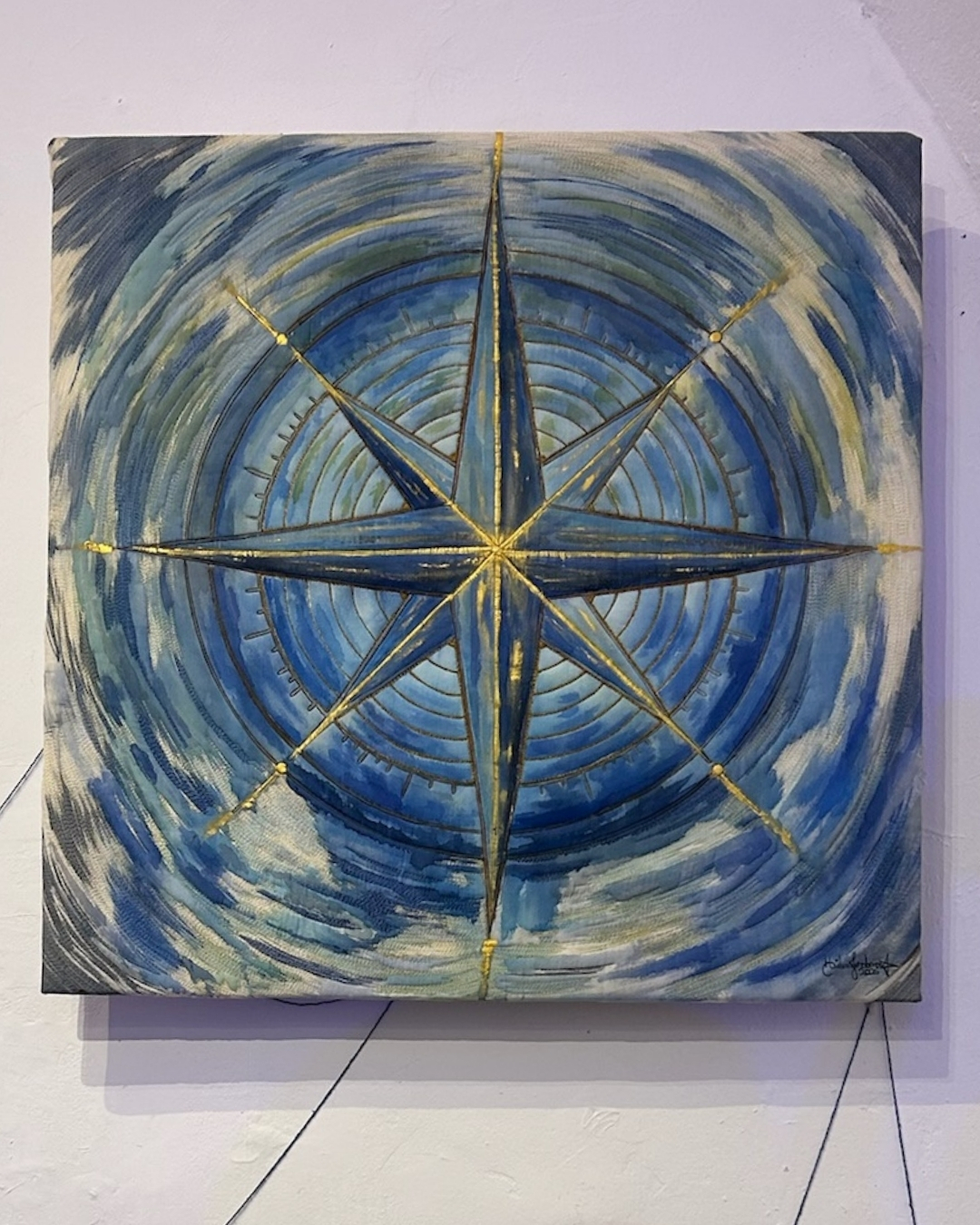 Obra de arte têxtil contemporânea intitulada "Minha Direção", de autoria de Hélen Jordana. A peça, de formato quadrado, é fixada em uma parede branca e apresenta uma imponente Rosa dos Ventos centralizada, desenvolvida com as técnicas de quilting e aquarela. O fundo e os círculos concêntricos são trabalhados em uma rica paleta de azuis, alternando tons profundos e claros com pinceladas fluidas e circulares que remetem ao movimento do mar e do vento. As linhas estruturais da Rosa dos Ventos, que formam a estrela de pontas e marcam as direções, são bordadas em relevo com linhas douradas reluzentes que irradiam a partir do centro exato da composição. Pequenos pontos dourados ornamentam os eixos. No canto inferior direito, nota-se a assinatura sutil da artista. Na base da imagem, aparecem fios pretos finos que fazem parte da expografia da instalação.
