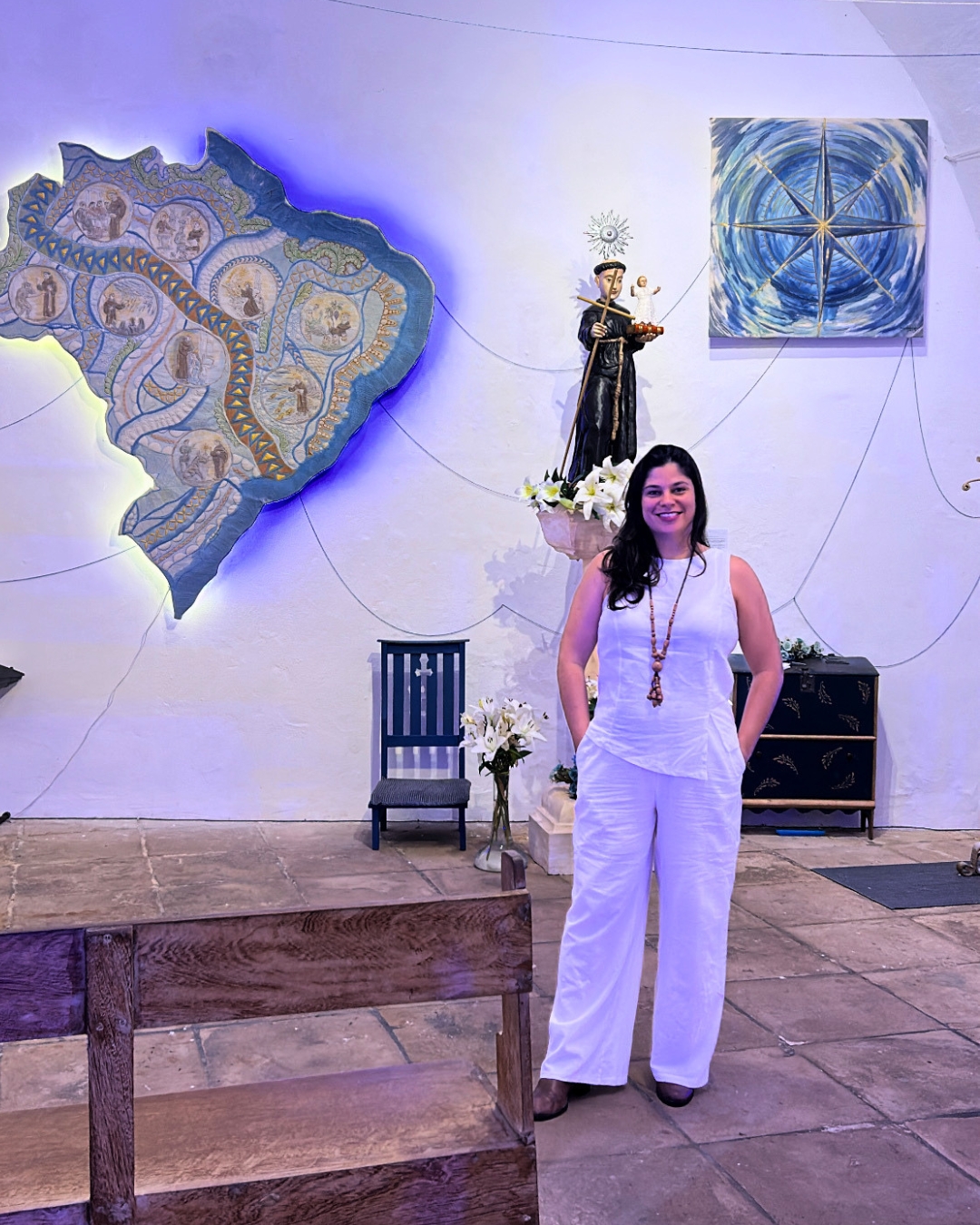 Fotografia da artista têxtil Hélen Jordana em pé na Sala de Exposições Temporárias do Forte Santo Antônio da Barra, posando sorridente à frente do Altar da Trezena de Santo Antônio. Hélen, uma mulher de cabelos longos e escuros, veste um conjunto todo branco de blusa sem mangas e calça pantalona, complementado por um colar comprido de contas. Ao fundo, na parede branca iluminada, estão expostas as suas duas obras de arte têxtil: à esquerda, o grande mapa do Brasil recortado em linho e acolchoado com quilting, iluminado por trás com uma luz azul suave; à direita, na parte superior, a obra "Minha Direção", uma tela quadrada com uma Rosa dos Ventos pintada em tons de azul e dourado. Entre as duas obras, sobre um pedestal decorado com lírios brancos, ergue-se a imagem escultural de Santo Antônio. Linhas finas conectam as obras e a imagem do santo na parede, criando uma ambientação poética. Na base da cena, avistam-se bancos de madeira fina e o piso rústico de pedra do forte.