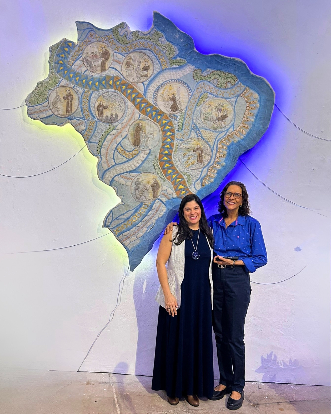 Fotografia de divulgação das artistas Irlânia Mercês e Hélen Jordana posando lado a lado na Sala de Exposições Temporárias do Forte Santo Antônio da Barra. À esquerda, a artista têxtil Hélen Jordana sorri vestindo um longo vestido azul-escuro, um colete longo bege claro e um colar comprido com pingente circular metálico. À direita, a artista de arte sacra Irlânia Mercês, usando óculos, sorri vestindo uma blusa azul de botões com pequenos detalhes rosados e calça escura. Ao fundo, centralizada na parede branca da galeria, destaca-se a imponente obra têxtil "Caminhos de Fé" de Hélen Jordana, um grande mapa do Brasil trabalhado em quilting e aquarela, iluminado por trás com uma luz azul e amarela suave que destaca seus contornos e os medalhões com os milagres de Santo Antônio. Finos fios pretos expográficos decoram a parede ao redor.