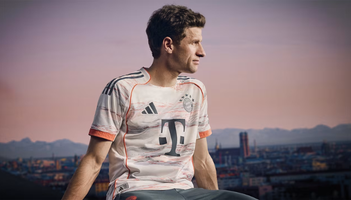 Camisa reserva do Bayern de Munique 2025-2026 branca com detalhes em cinza e laranja, com escudo bordado e homenagem à Allianz Arena.