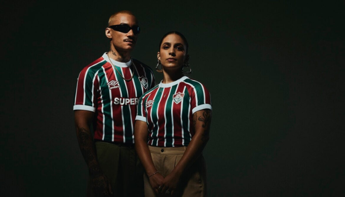 Camisa oficial Fluminense 2025-2026, modelo titular, com listras verticais em vermelho, branco e gren&aacute;, gola redonda branca e detalhes em gren&aacute; na nuca. Escudo do Fluminense bordado no lado esquerdo do peito, logo da Umbro em branco com bordas gren&aacute;s no lado direito e patroc&iacute;nio Superbet centralizado. Etiqueta na barra frontal com a frase &ldquo;Das Laranjeiras para o Mundo&rdquo;, em refer&ecirc;ncia &agrave; participa&ccedil;&atilde;o no Mundial.