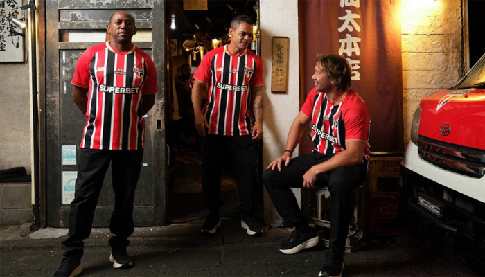 Imagem da camisa reserva do São Paulo FC 2025-2026, com listras vermelhas e pretas, gola V vermelha e detalhes dourados nos punhos.