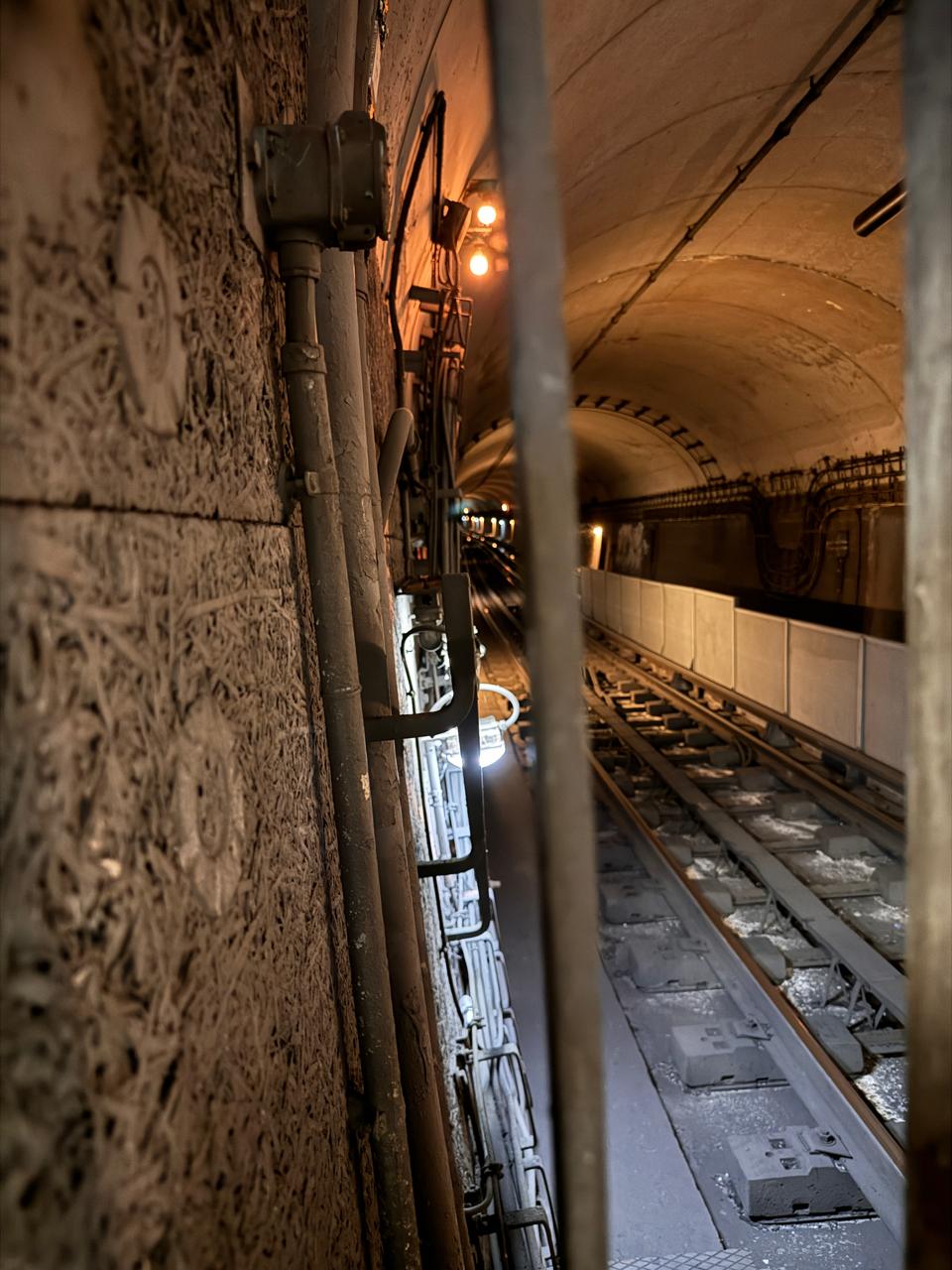 Homem de costas (Prosa Graffiti) olhando para o horizonte de um túnel ferroviário escuro em Paris, segurando latas de spray, com foco na perspectiva dos trilhos.