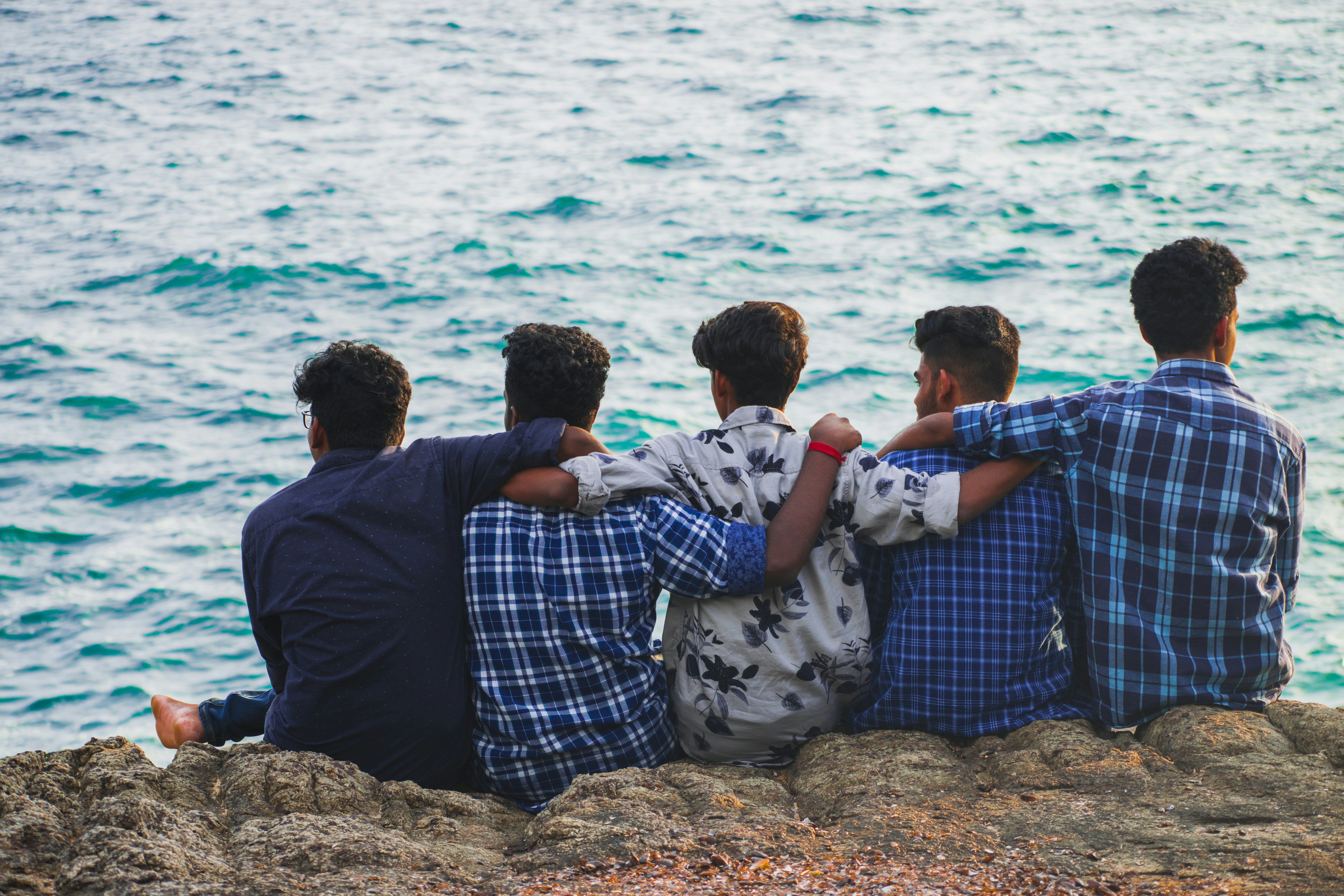 Cinco amigos sentados en la costa con los brazos entrelazados, mirando el mar, compartiendo un momento de conexión y camaradería.