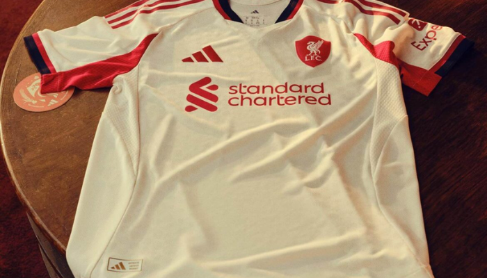 Camisa reserva Liverpool Adidas 2025/26 off-white com gola V, três listras vermelhas e escudo inspirado em Anfield 1906.