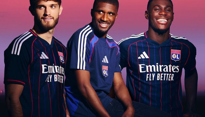 Camisa Reserva Lyon Adidas 2025-26 azul marinho com linhas azul royal e vermelhas, gola Y, três listras prateadas e frase “Orgulhosos de nossas cores” no interior da gola.