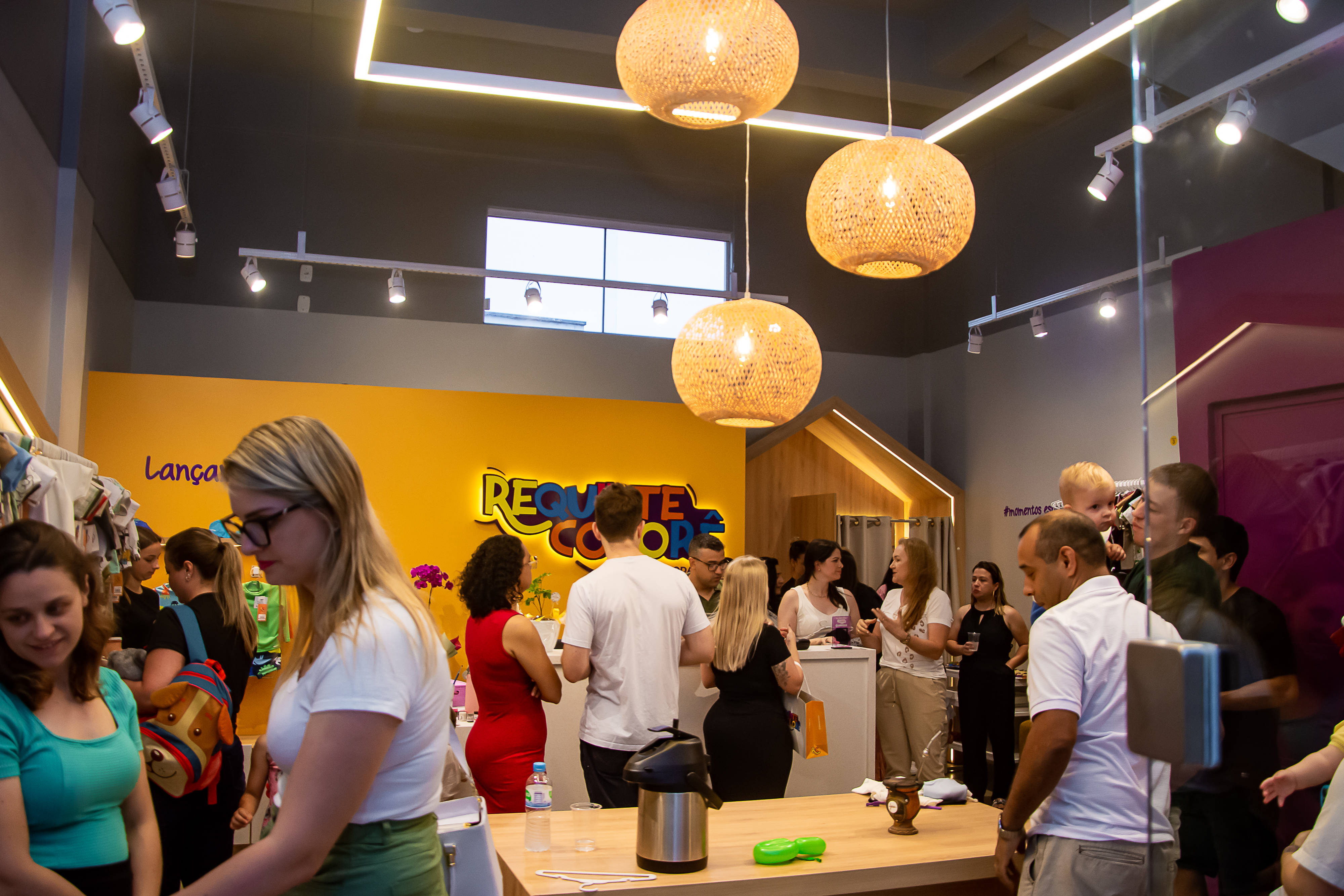 Interior da loja Requinte Colorê com araras de roupas infantis e pessoas reunidas no espaço.
