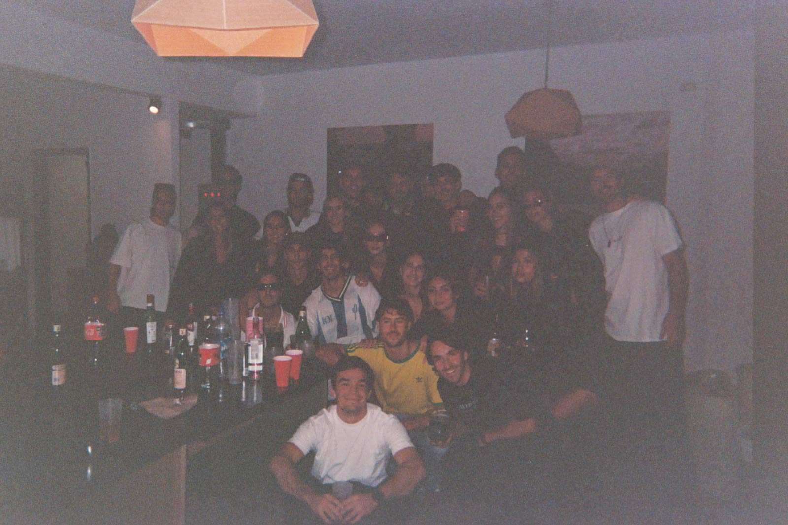 Foto analógica de un encuentro en House of Tyrol: un grupo numeroso de amigos reunidos alrededor de la barra, con vasos y botellas.