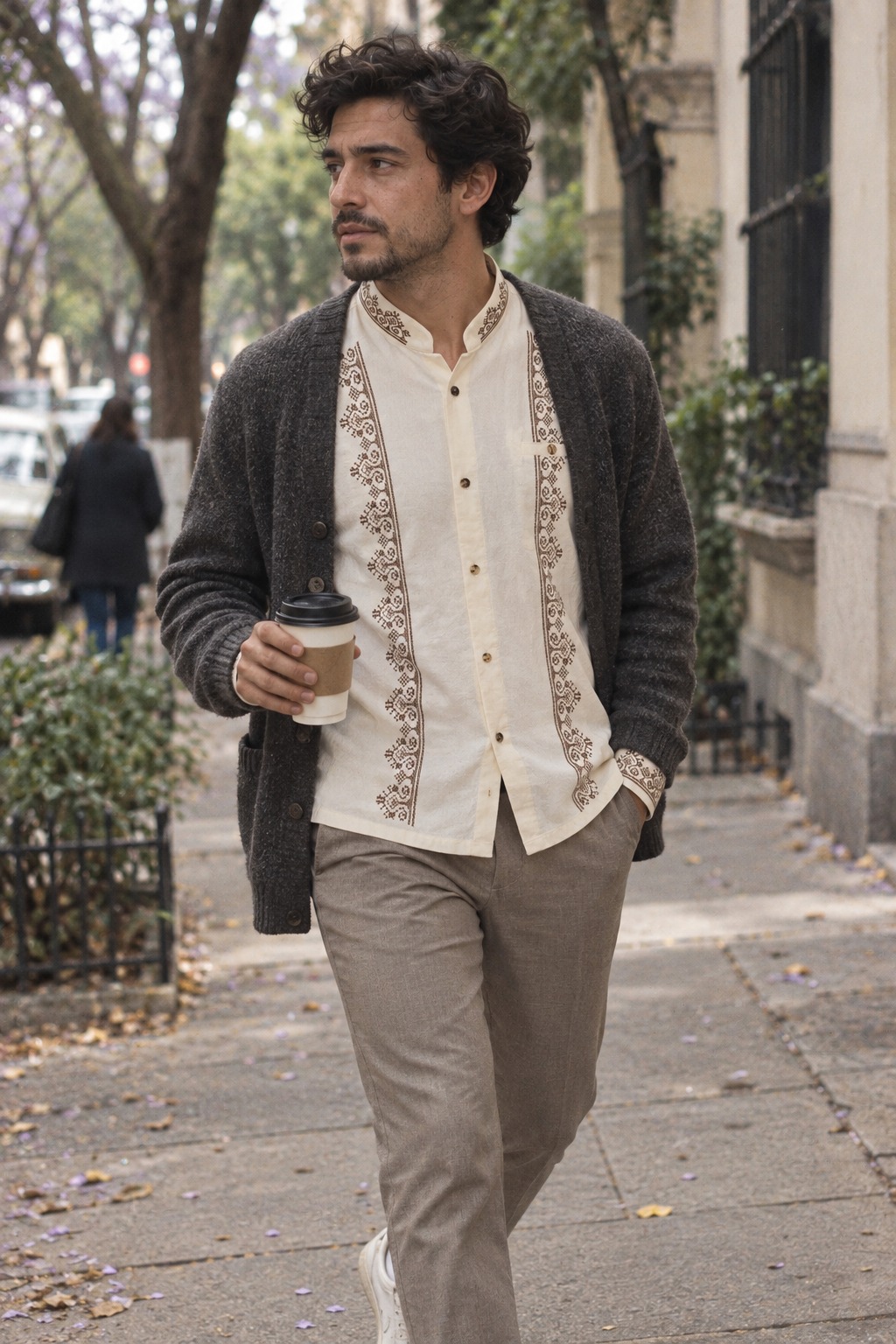 Hombre con camisa Tizil beige-crudo con bordados caf&eacute; bajo un c&aacute;rdigan gris oscuro, con un caf&eacute; en la mano, en una calle arbolada de la Ciudad de M&eacute;xico