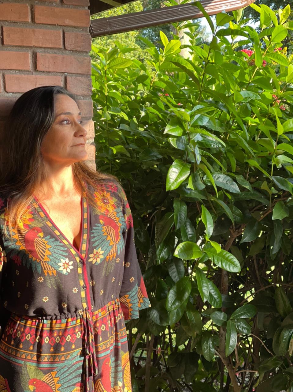 Mulher branco com cabelo castanhos, vestido estampado, sob a luz do sol, encostada na parede e olhando para o horizonte.