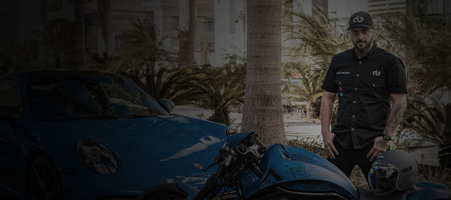 Homem com barba e tatuagens usando camisa preta com patches e boné da DB Studio Garage, posando ao lado de uma motocicleta custom azul e um carro esportivo Porsche azul, em ambiente externo com palmeiras ao fundo.