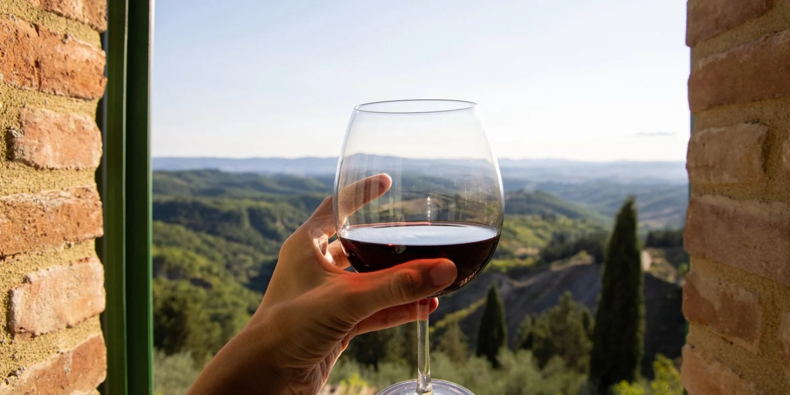 Ta&ccedil;a de vinho tinto diante de uma paisagem de colinas, enquadrada por uma janela r&uacute;stica.