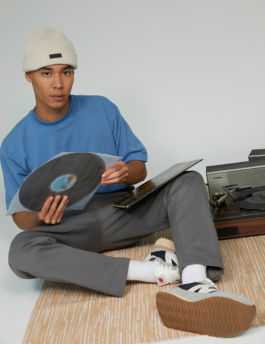 Modelo vestindo roupas da ikiwa segurando um disco vinil antigo e sentado em um tapete claro ao lado de uma vitrola
