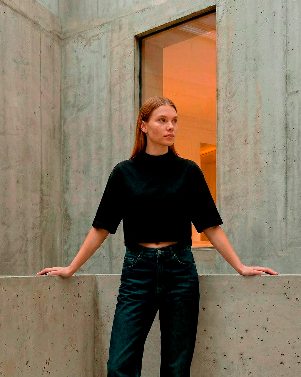 Uma mulher jovem de pele clara e cabelos longos e lisos posa em um ambiente de arquitetura minimalista com paredes de concreto aparente. Ela veste uma camiseta cropped preta de gola alta da ikiwa, com mangas que vão até o cotovelo, e uma calça jeans de lavagem escura e cintura alta. Ela está apoiada em uma mureta de concreto, com os braços abertos para as laterais. Ao fundo, uma janela vertical estreita revela uma luz interna quente e alaranjada, contrastando com o tom frio e industrial das paredes de cimento.