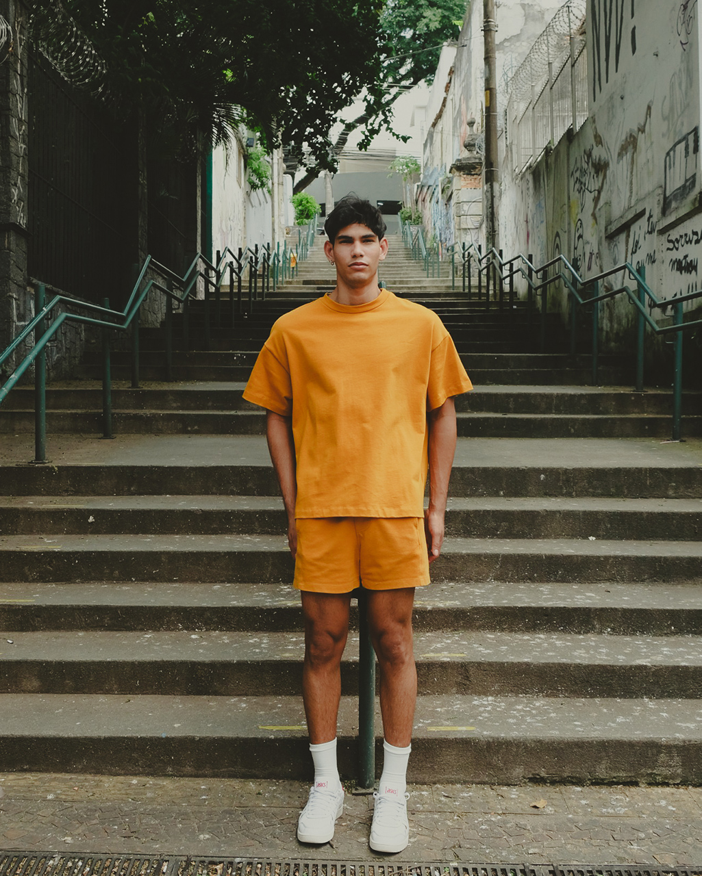 Um homem jovem de pele clara e cabelos escuros curtos está em pé, centralizado em uma escadaria de pedra ao ar livre. Ele veste um conjunto monocromático na cor açafrão, composto por uma camiseta de modelagem Boxy da ikiwa com gola encorpada e um shorts acima do joelho de mesmo tom e modelagem Up Cut da ikiwa. Ele usa meias brancas de cano alto e tênis brancos. O fundo apresenta uma escadaria longa que sobe entre muros com grafites discretos e árvores ao topo. A iluminação é suave e levemente amarelada, conferindo um aspecto urbano e moderno à fotografia.