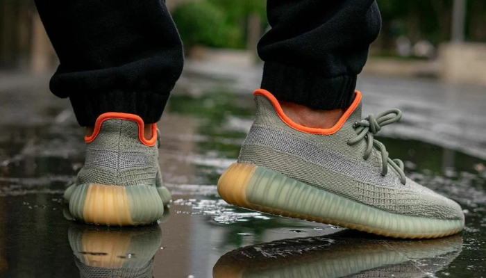 Tênis Yeezy Boost 350 V2 Desert Sage em Primeknit verde claro com detalhes laranja e faixa reflexiva.