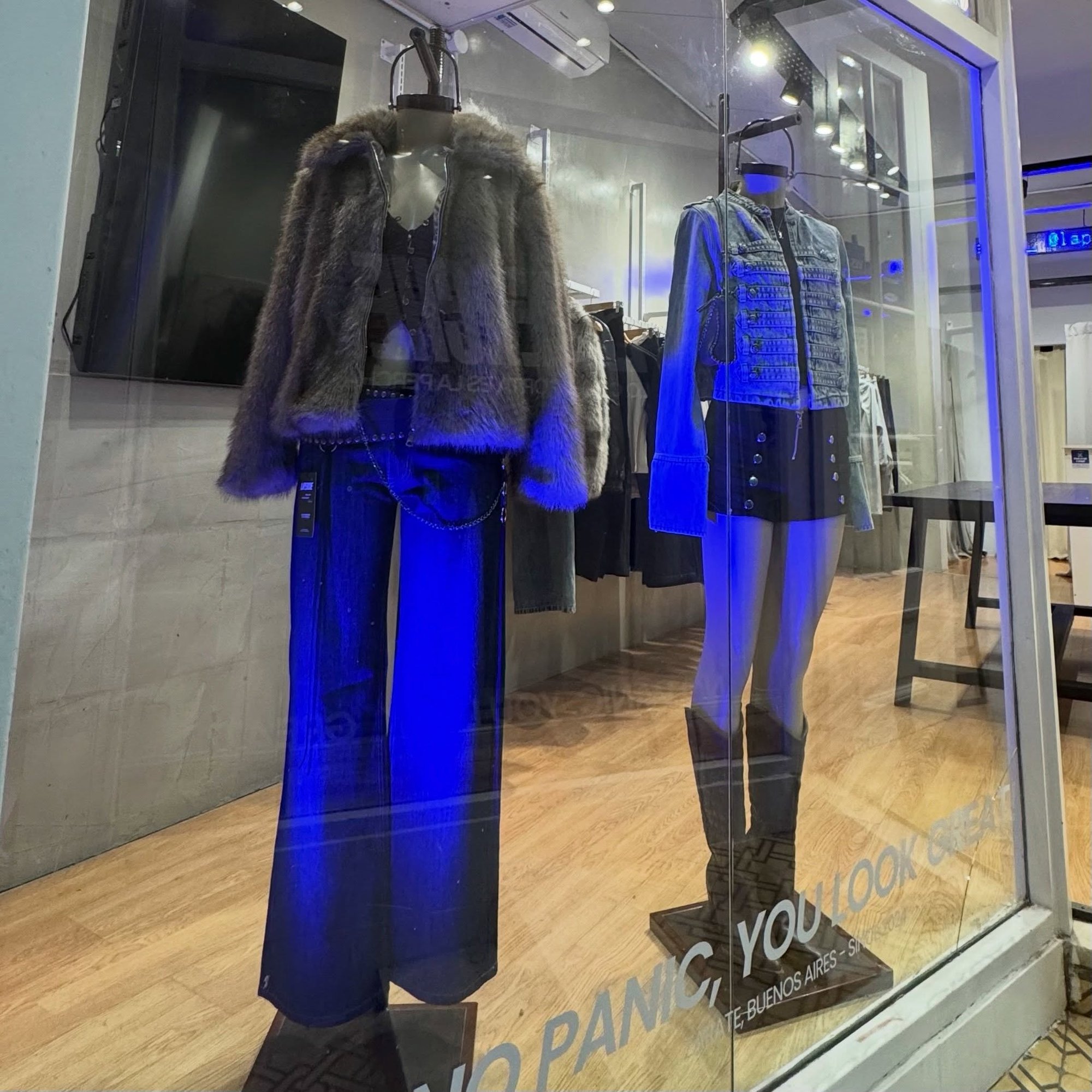 Vidriera de La Perche de noche, con dos maniquíes vestidos y el cartel NO PANIC, YOU LOOK GREAT sobre el vidrio