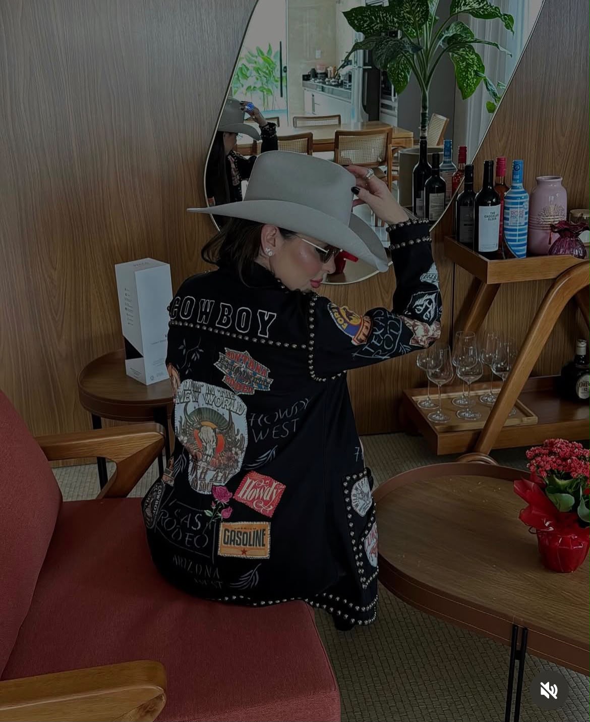 Mari Perillo de costas, com jaqueta preta bordada com patches e chapéu cinza
