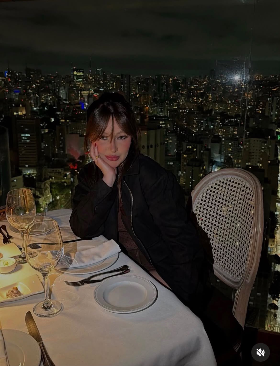 Mari Perillo à mesa de um restaurante, com a cidade iluminada ao fundo