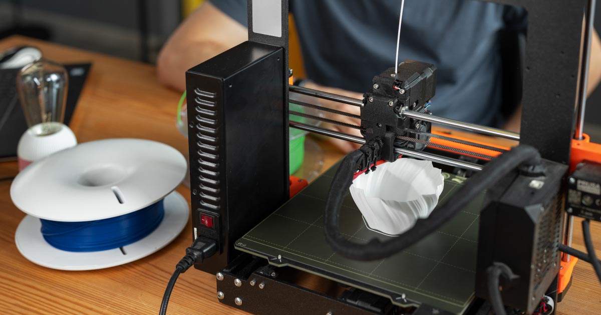 Impressora 3D em funcionamento imprimindo objeto branco, com filamento azul ao lado, sobre mesa de madeira.