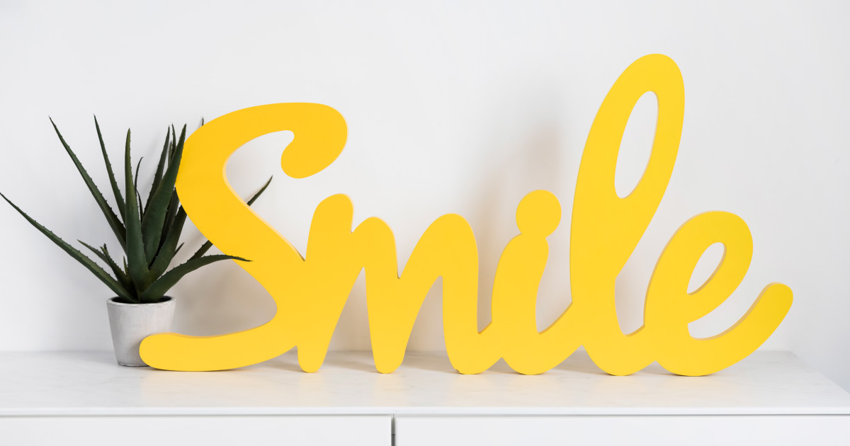Letreiro decorativo amarelo com a palavra 'Smile' em 3D ao lado de vaso com planta sobre superfície branca.