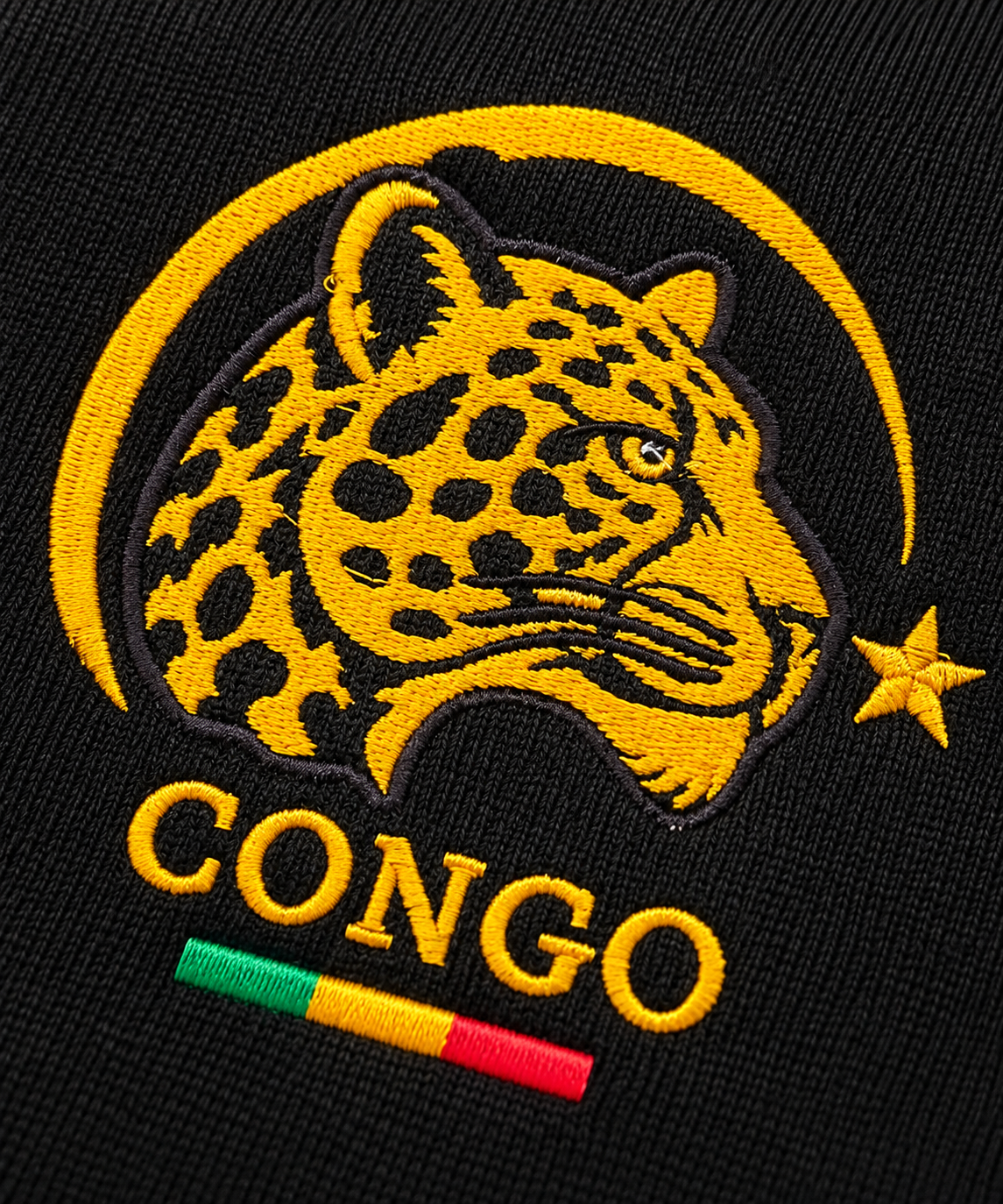 Detalhe do emblema do leopardo bordado em amarelo sobre a malha preta da camisa Congo Heritage
