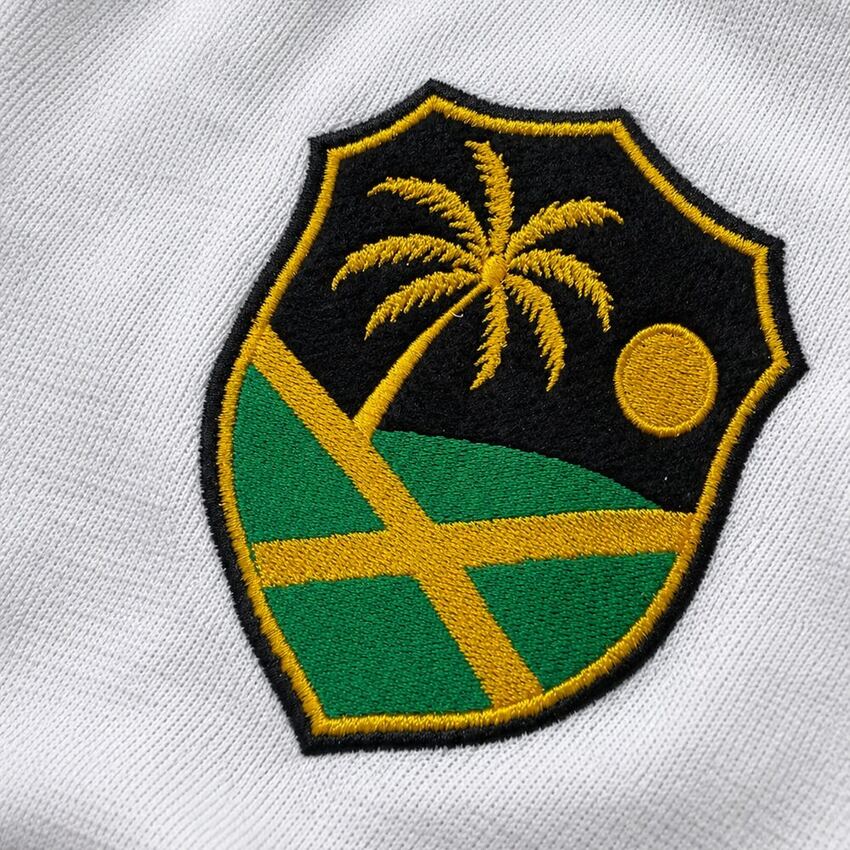 Close macro do bras&atilde;o bordado: palmeira, sol e formas da bandeira jamaicana sobre tecido branco